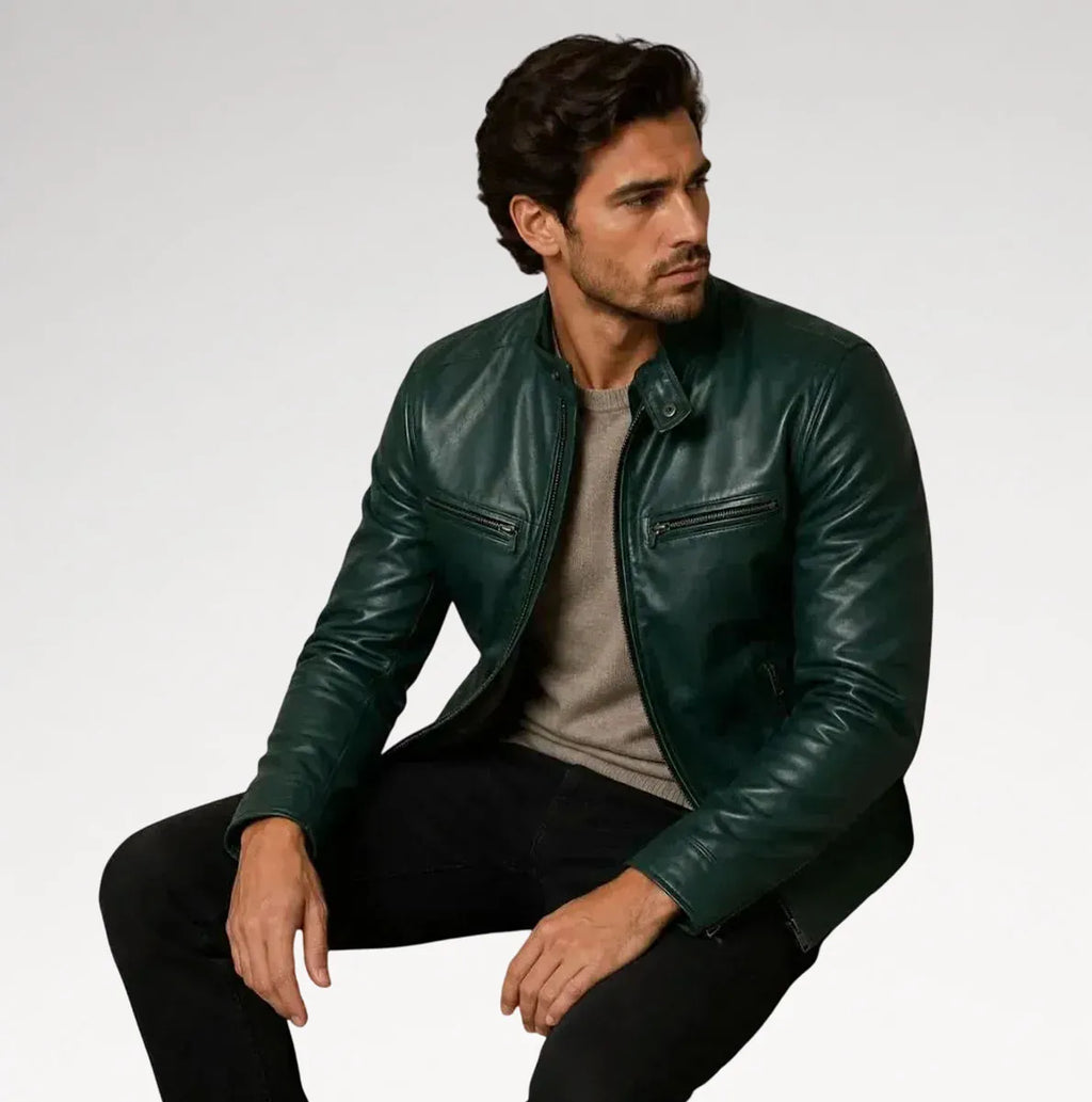 A man wearing a dark green leather jacket over a gray t-shirt and black pants, sitting against a light gray studio background.