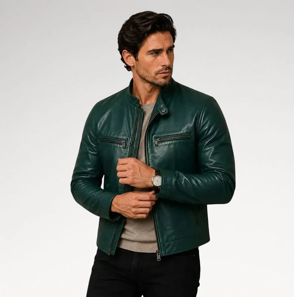 A man wearing a dark green leather jacket over a light gray t-shirt and black pants, standing against a light gray studio background.