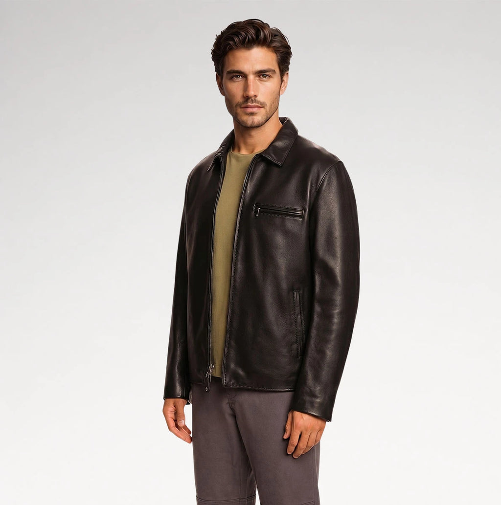 A man wearing a dark brown leather jacket over an olive green shirt and gray pants, standing against a light gray studio background.