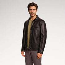A man wearing a dark brown leather jacket over an olive green shirt and gray pants, standing against a light gray studio background.