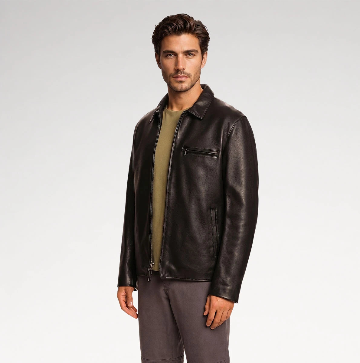 A man wearing a dark brown leather jacket over an olive green shirt and gray pants, standing against a light gray studio background.