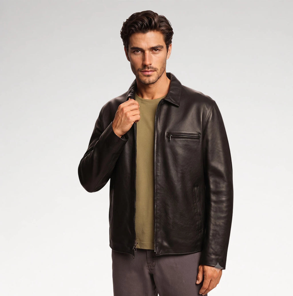 A man wearing a dark brown leather jacket over an olive green t-shirt and gray pants, standing against a light gray studio background.