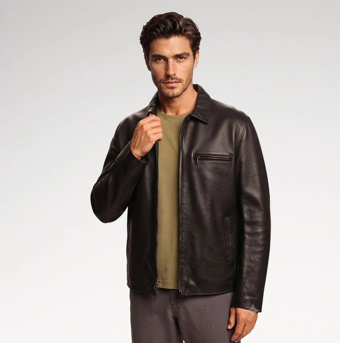 A man wearing a dark brown leather jacket over an olive green t-shirt and gray pants, standing against a light gray studio background.