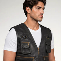 A man wearing a black leather utility vest over a white t-shirt, shown from the chest up against a light gray studio background.