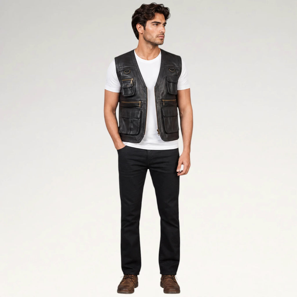A man modeling a black leather utility vest with multiple front pockets, worn over a white t-shirt and black jeans, standing against a light gray studio background.