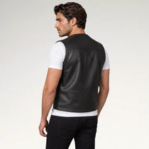 A man wearing a black leather vest over a white t-shirt and black pants, shown from a three-quarters back view against a light gray studio background.