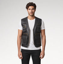 A man modeling a black leather utility vest with multiple zippered and buttoned pockets over a white t-shirt and black pants, shot against a light gray studio background.