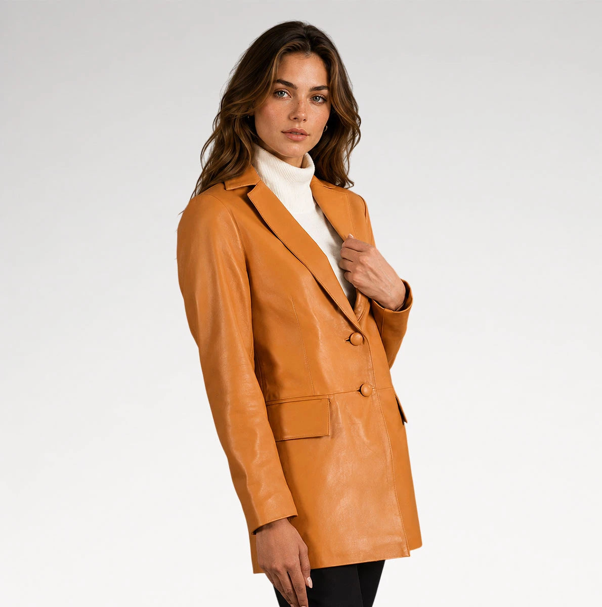 A woman wearing a tan leather blazer over a white turtleneck sweater and black pants, photographed against a clean light gray studio background.