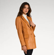 A woman modeling a tan faux-leather blazer over a white turtleneck sweater and black pants, photographed against a light gray studio background.