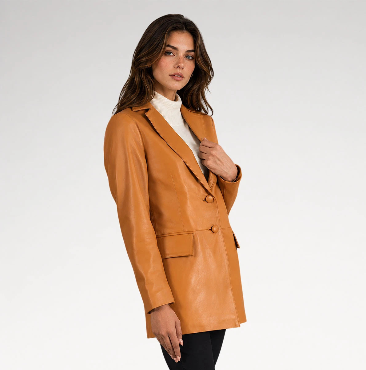 A woman modeling a tan faux-leather blazer over a white turtleneck sweater and black pants, photographed against a light gray studio background.