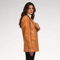 A woman wearing a camel brown leather blazer over a white turtleneck and black pants, photographed from the side against a light gray studio background.