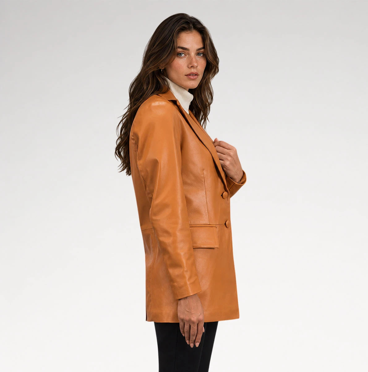A woman wearing a camel brown leather blazer over a white turtleneck and black pants, photographed from the side against a light gray studio background.