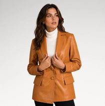 A woman wearing a camel brown faux leather blazer over a white turtleneck sweater and black pants, standing against a light gray studio background.