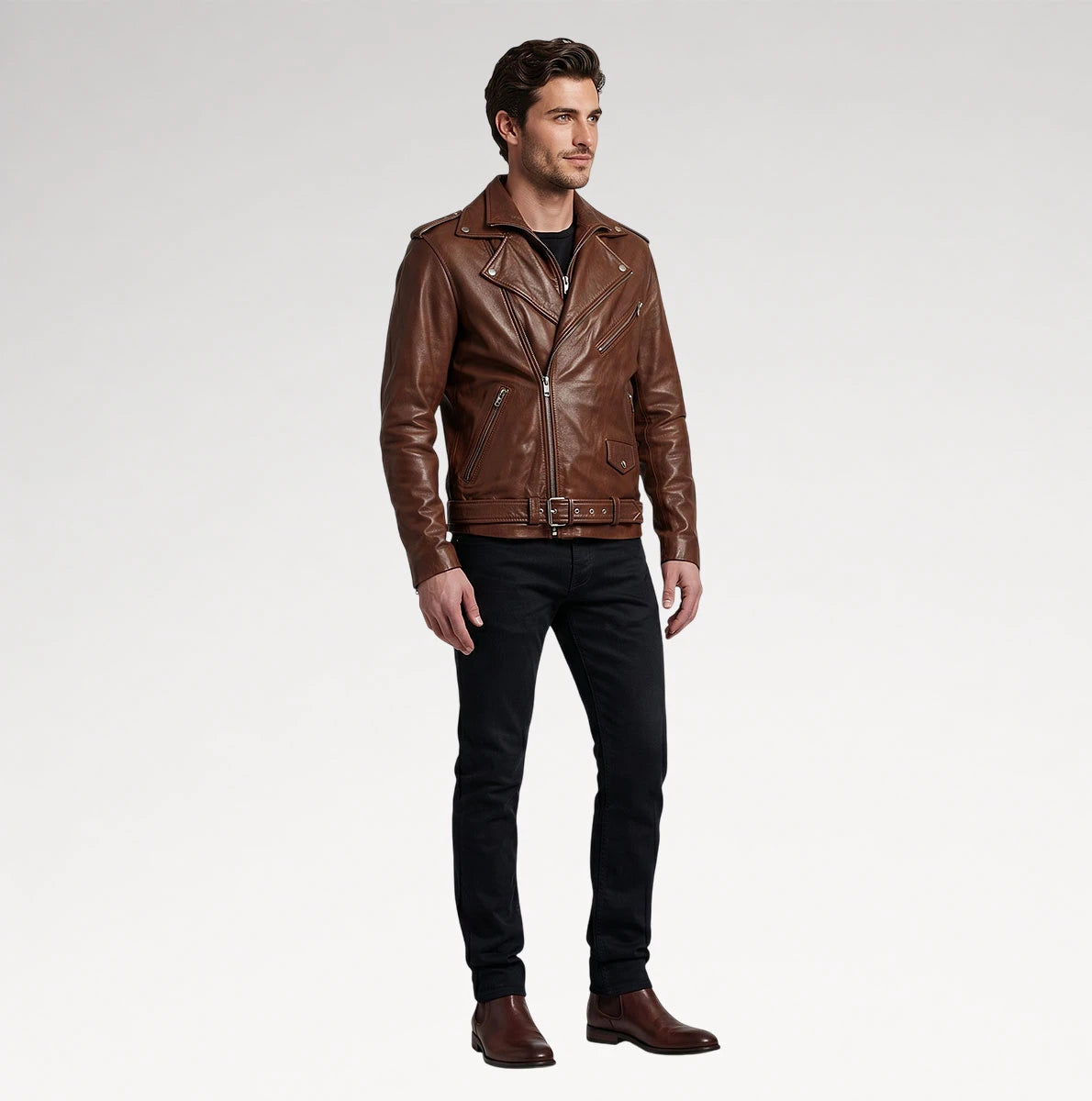 A man modeling a brown leather moto jacket, paired with black jeans and brown boots, standing against a light gray studio background.
