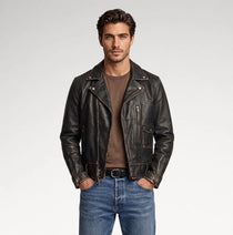 A man wearing a black distressed leather motorcycle jacket over a brown t-shirt and blue jeans, standing against a light gray studio background.