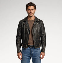 A man wearing a black leather motorcycle jacket over a brown t-shirt and blue jeans, standing against a light gray studio background.
