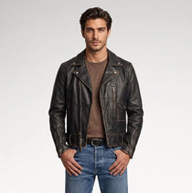 A man wearing a black distressed leather motorcycle jacket over a brown t-shirt and blue jeans, standing against a light gray studio background.