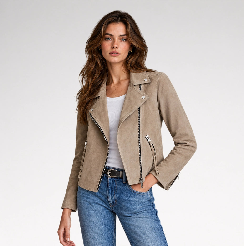 A woman modeling a taupe suede moto jacket over a white tank top and blue jeans, photographed against a clean light gray studio background.