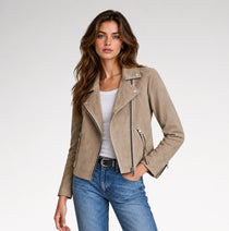 A woman modeling a taupe suede moto jacket over a white tank top and blue jeans, photographed against a clean light gray studio background.