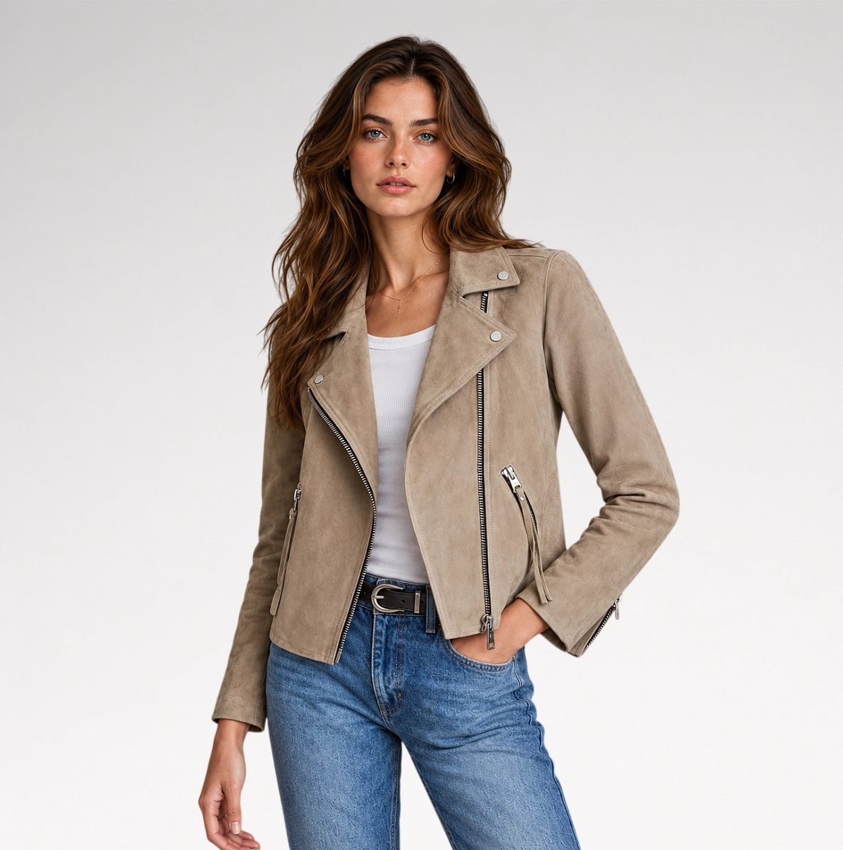 A woman modeling a taupe suede moto jacket over a white tank top and blue jeans, photographed against a clean light gray studio background.