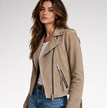 A woman wearing a beige suede moto jacket with silver zippers, styled with a white top and blue jeans, photographed against a clean white studio background.
