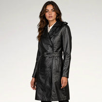 A woman modeling a black leather trench coat with a matching belt, photographed against a plain light gray studio background.