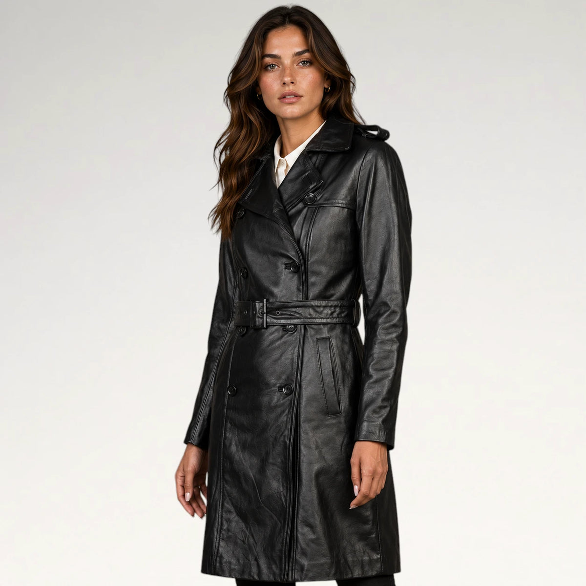 A woman modeling a black leather trench coat with a matching belt, photographed against a plain light gray studio background.