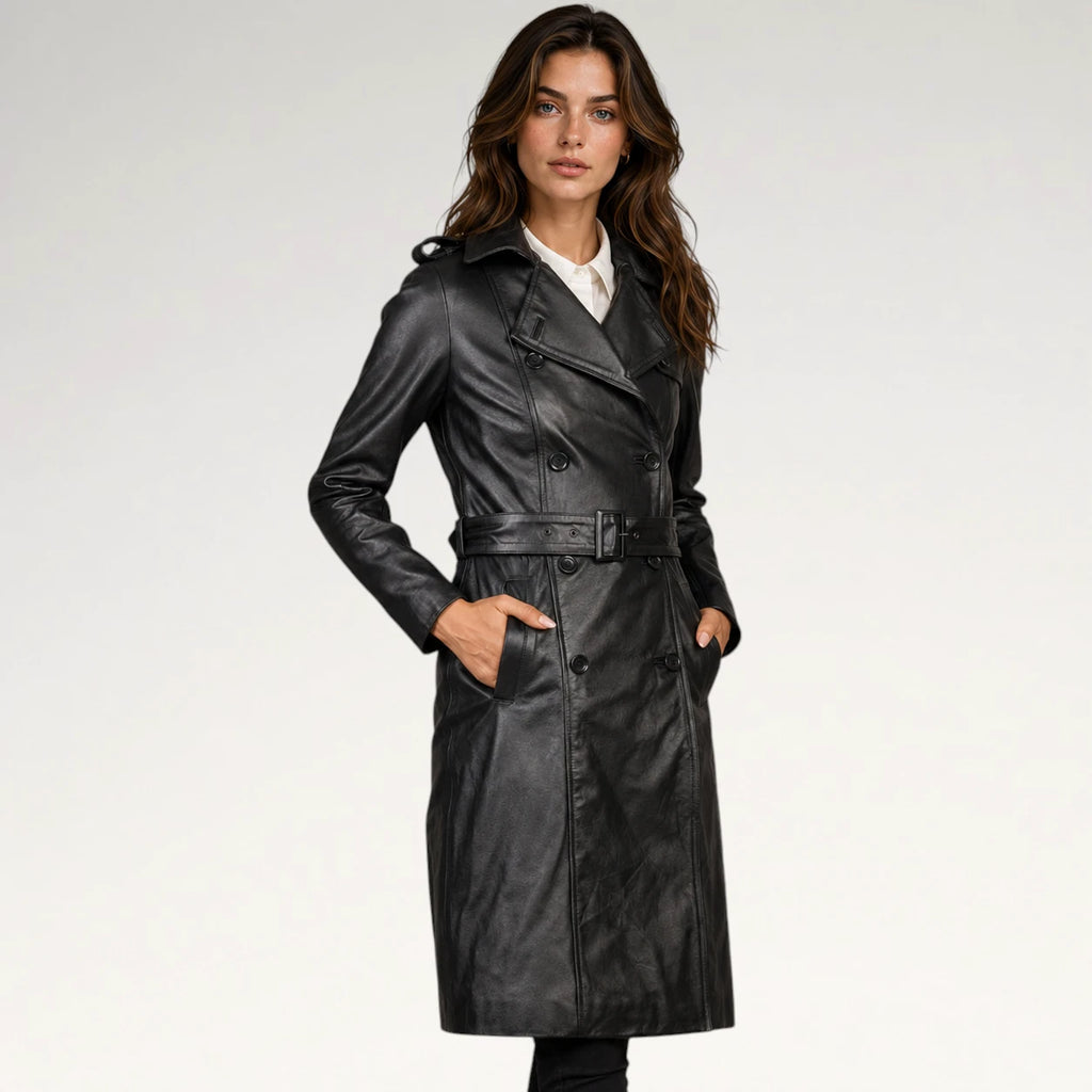 A woman modeling a black leather trench coat with a belted waist, double-breasted front, and wide lapels, photographed against a clean light gray studio background.
