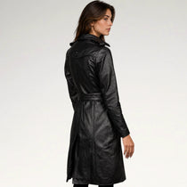 A woman wearing a black leather trench coat with a belted waist, shown from a three-quarters back view against a plain white studio background.