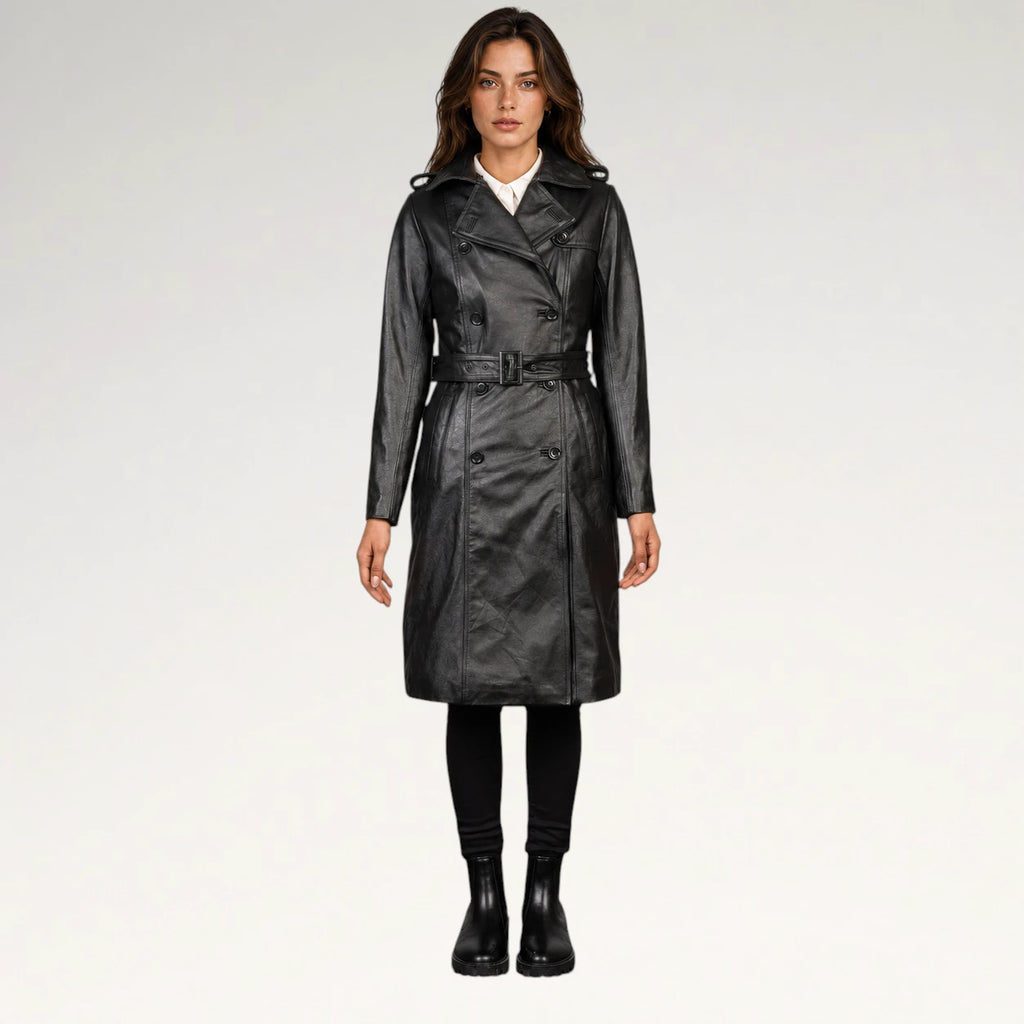 A woman modeling a black faux-leather trench coat with a matching belt, worn over a white shirt and black pants, against a plain light gray studio background.