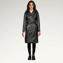 A woman modeling a black faux-leather trench coat with a matching belt, worn over a white shirt and black pants, against a plain light gray studio background.