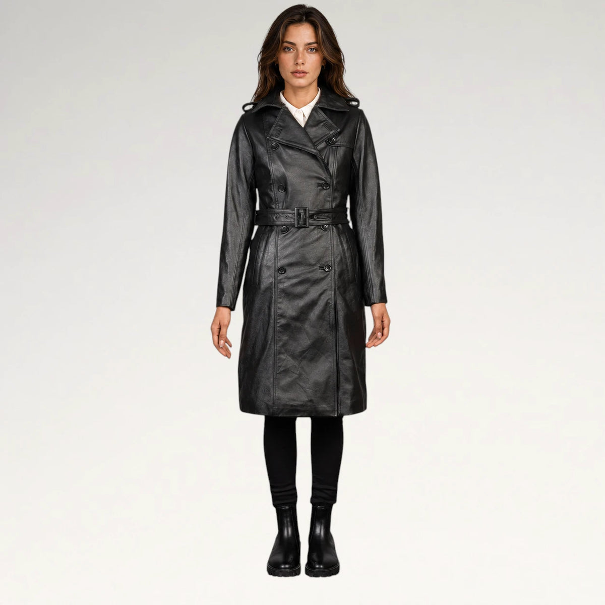 A woman modeling a black faux-leather trench coat with a matching belt, worn over a white shirt and black pants, against a plain light gray studio background.