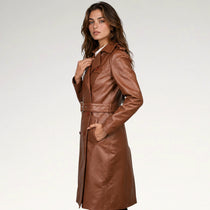A woman modeling a long brown faux-leather trench coat with a matching belt, shown against a plain light gray studio background.