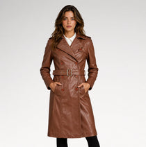 A woman modeling a brown faux-leather double-breasted trench coat with a matching belt, worn over a white collared shirt and black pants, against a light gray studio background.