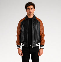 A man wearing a black and brown leather varsity-style jacket with white striped ribbed cuffs and collar, standing against a light gray studio background.