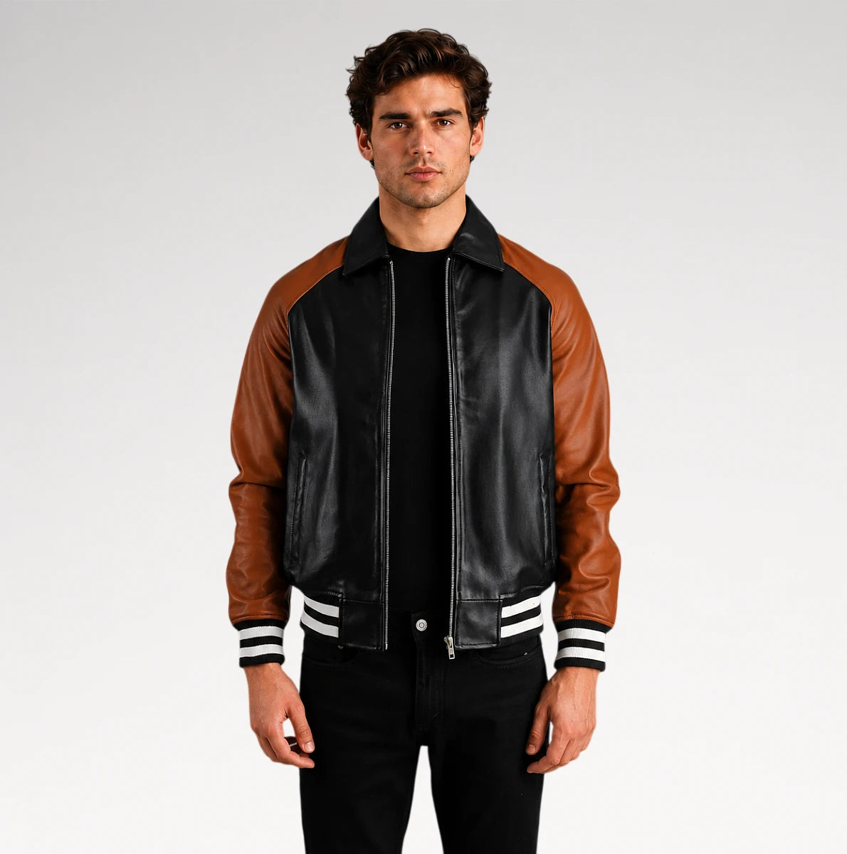 A man wearing a black and brown leather varsity-style jacket with white striped ribbed cuffs and collar, standing against a light gray studio background.