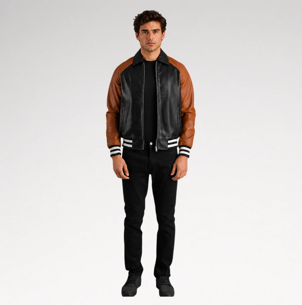 A man modeling a black leather and brown faux-leather varsity-style jacket with white striped ribbed cuffs and collar, paired with black pants and black shoes, against a light gray studio background.
