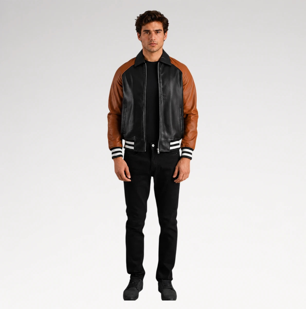 A man modeling a black leather and brown faux-leather varsity-style jacket with white striped ribbed cuffs and collar, paired with black pants and black shoes, against a light gray studio background.