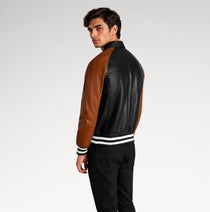 A man wearing a black and brown leather varsity-style jacket with white striped ribbed cuffs and hem, shown from a side-back angle against a light gray studio background.