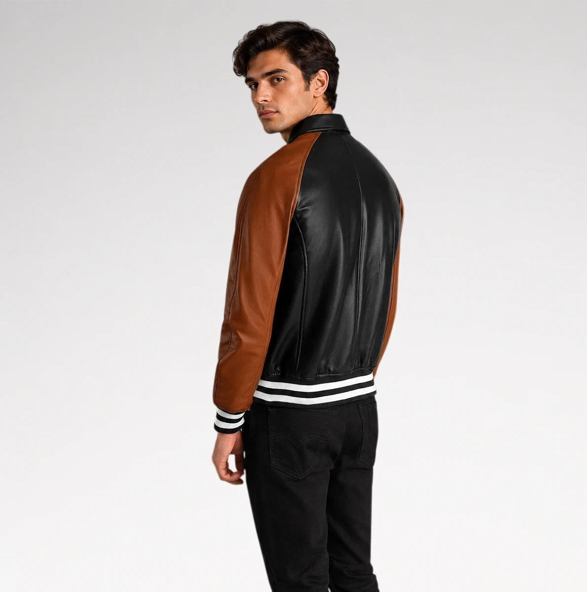 A man wearing a black and brown leather varsity-style jacket with white striped ribbed cuffs and hem, shown from a side-back angle against a light gray studio background.