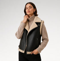 A woman models a black faux-leather moto jacket with a beige sherpa collar and lining, worn over a beige ribbed turtleneck sweater and black pants, against a light gray studio background.