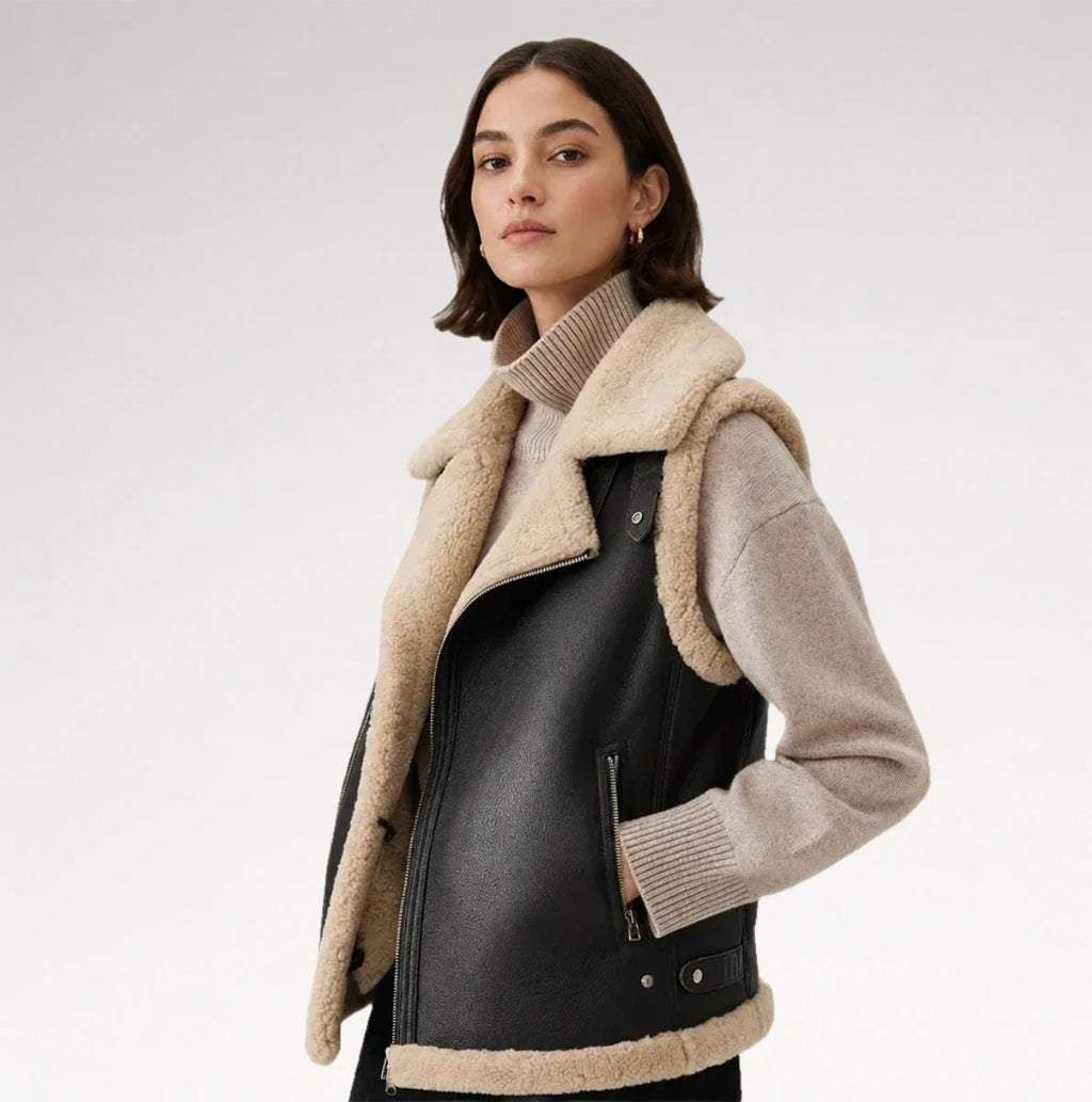 A woman models a black faux-leather moto vest with beige shearling lining and knit ribbed turtleneck and cuff inserts, photographed against a clean light gray studio background.