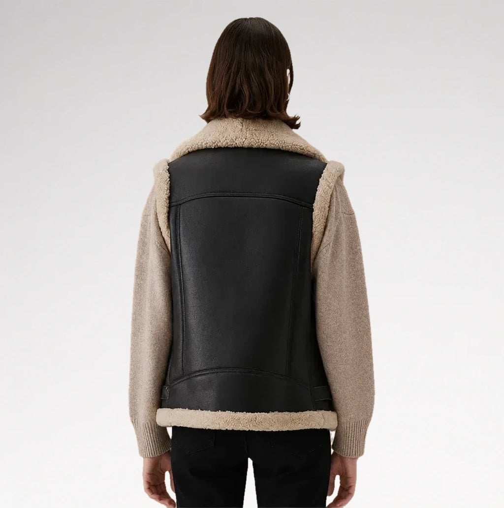 A model shown from the back wearing a black faux-leather vest with beige shearling lining and knit sleeves, paired with black pants, against a light gray studio background.