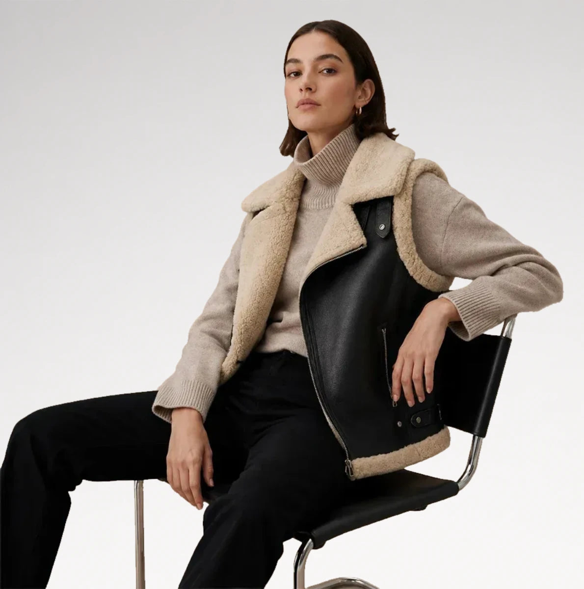 A woman seated on a black chair wearing a beige shearling-lined black leather jacket over a beige turtleneck sweater and black trousers, against a clean light gray studio background.