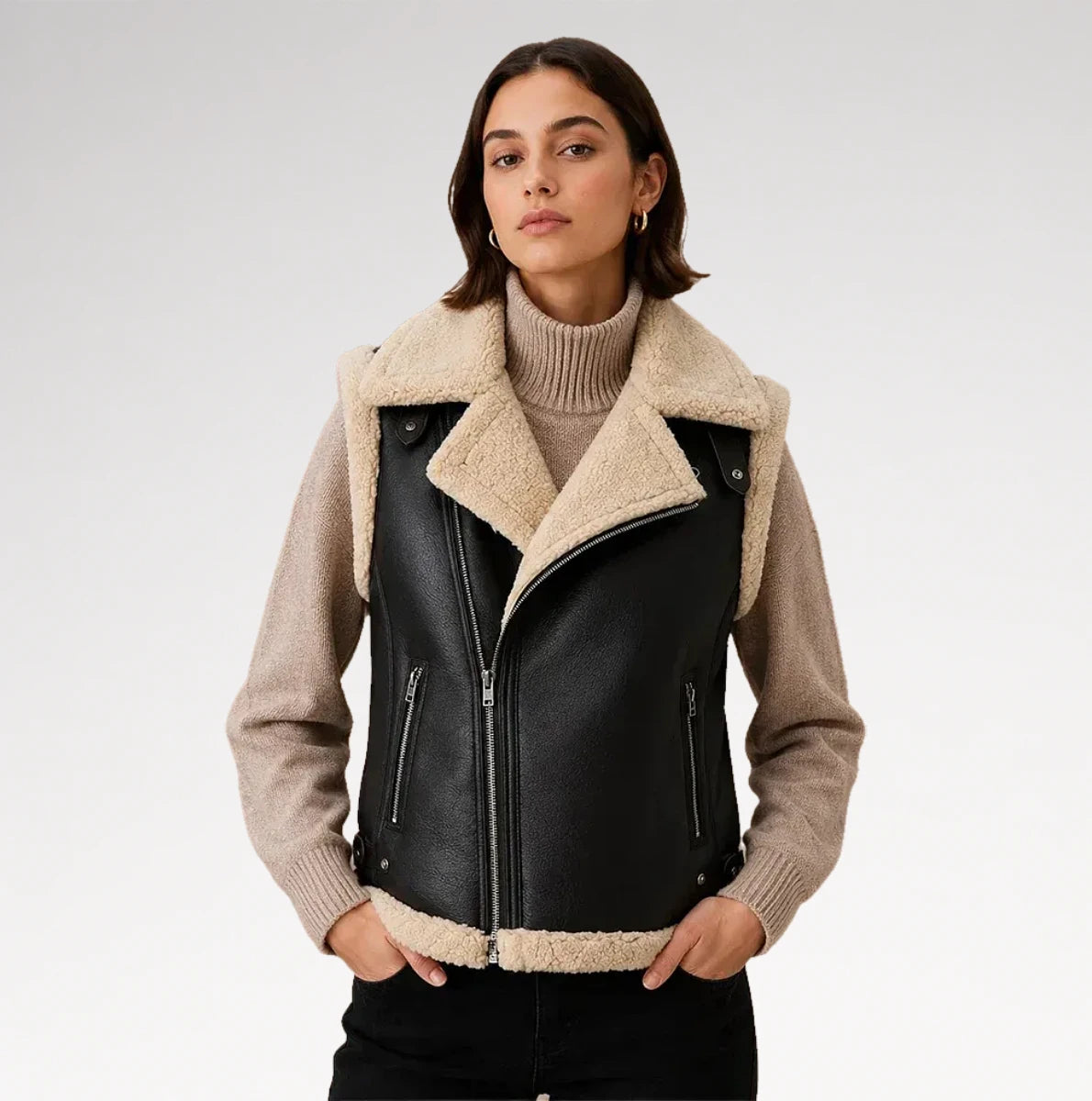A woman wearing a black faux-leather moto jacket with a beige sherpa collar and lining, layered over a beige ribbed turtleneck sweater and black pants, photographed against a light gray studio background.