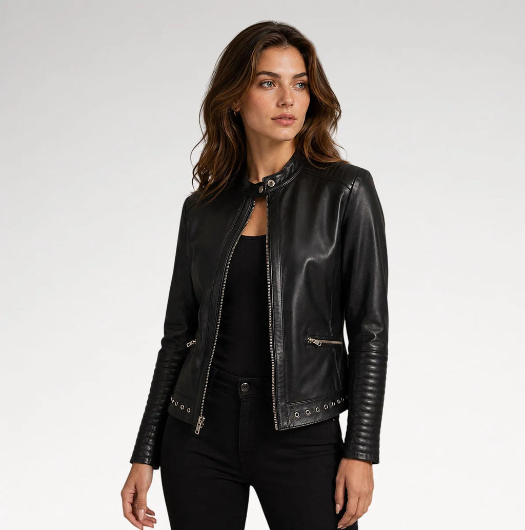 A woman modeling a black faux-leather moto jacket with silver-tone hardware, styled over a black top and black pants against a light gray studio background.