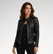 A woman modeling a black faux-leather moto jacket with silver-tone hardware, styled over a black top and black pants against a light gray studio background.