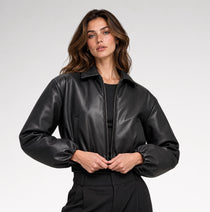 A woman wearing a black faux-leather bomber jacket with a stand-up collar and elasticized cuffs, posing against a light gray studio background.