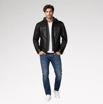 A man modeling a black leather jacket with a zippered hood, worn over a white t-shirt and blue jeans, on a light gray studio background.