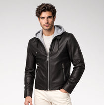 A man wearing a black leather zip-up hooded jacket with a gray fleece-lined hood, layered over a white t-shirt and cream pants, photographed against a light gray studio background.